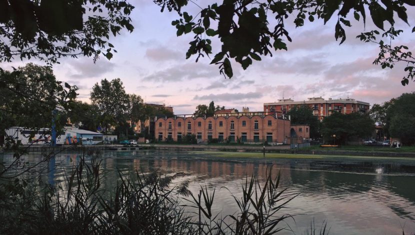 Un fiume in primo piano con alberi e vegetazione; sull'altra riva, un grande edificio in mattoni