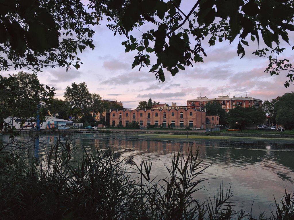 Un fiume in primo piano con alberi e vegetazione; sull'altra riva, un grande edificio in mattoni