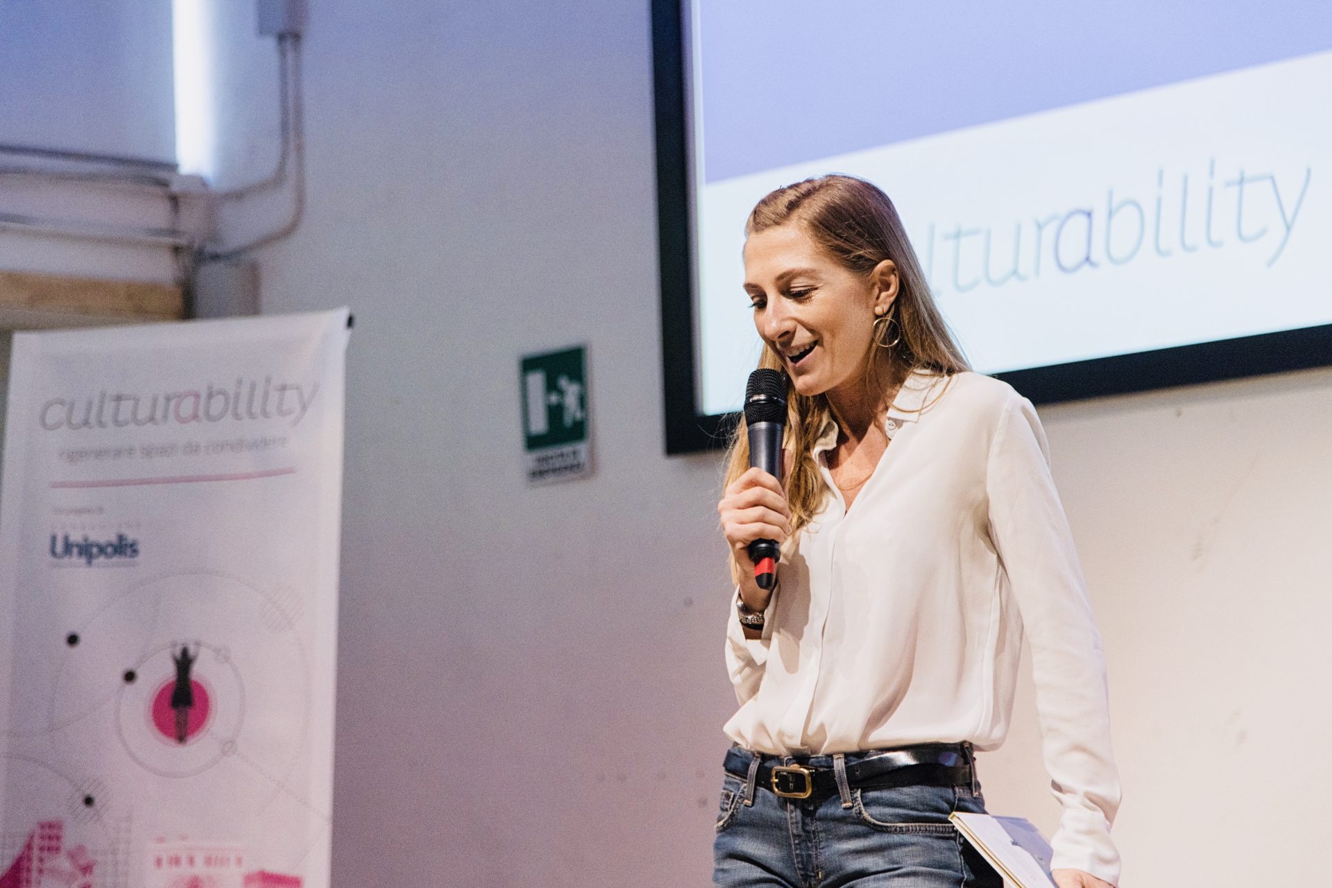 Roberta Franceschinelli in camicia bianca e jeans in piano americano, che parla a un microfono; sullo sfondo uno schermo