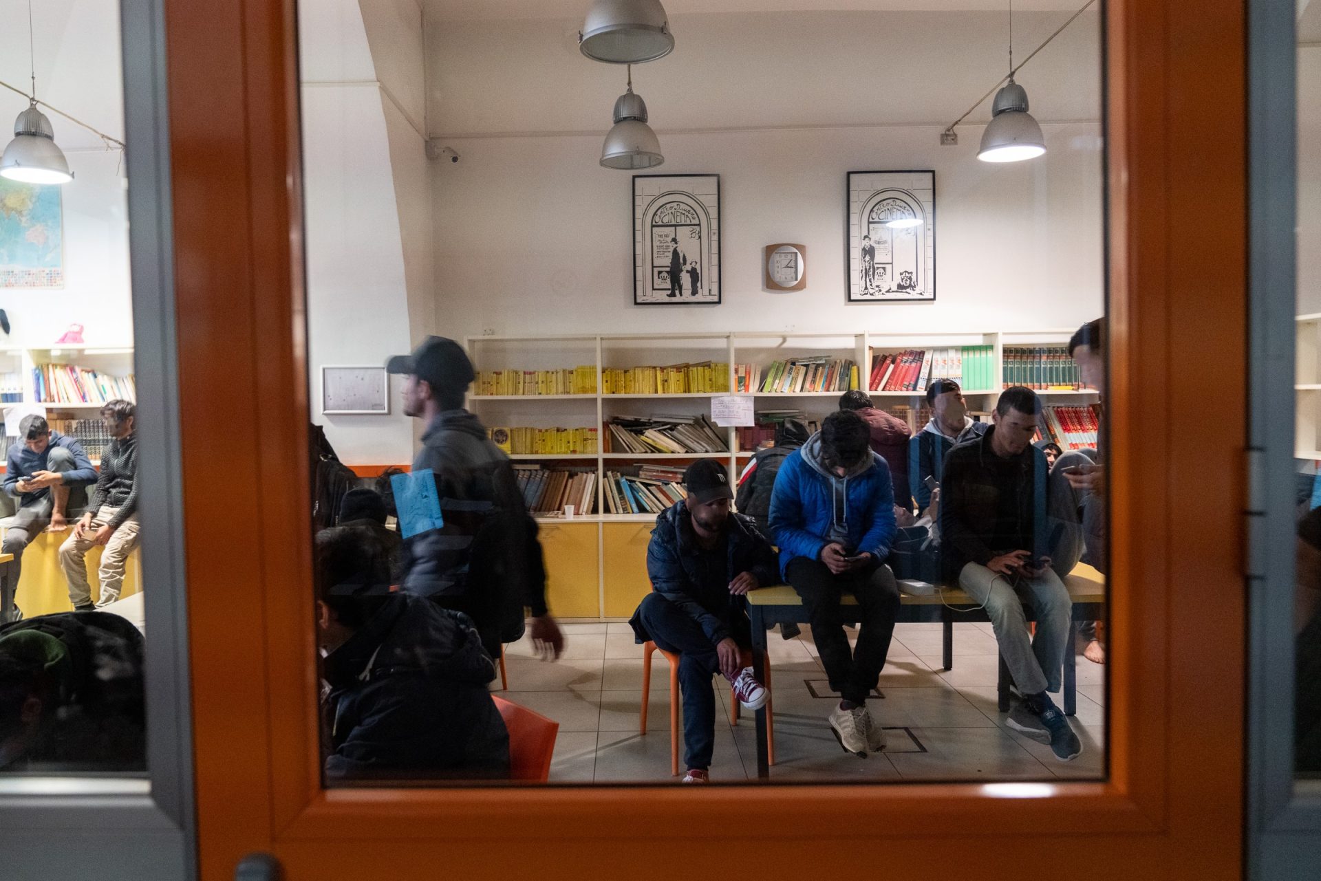 Attraverso un vetro, si vedono alcune persone nel centro diurno, sedute su un tavolo. Sullo sfondo, una libreria e dei quadri 