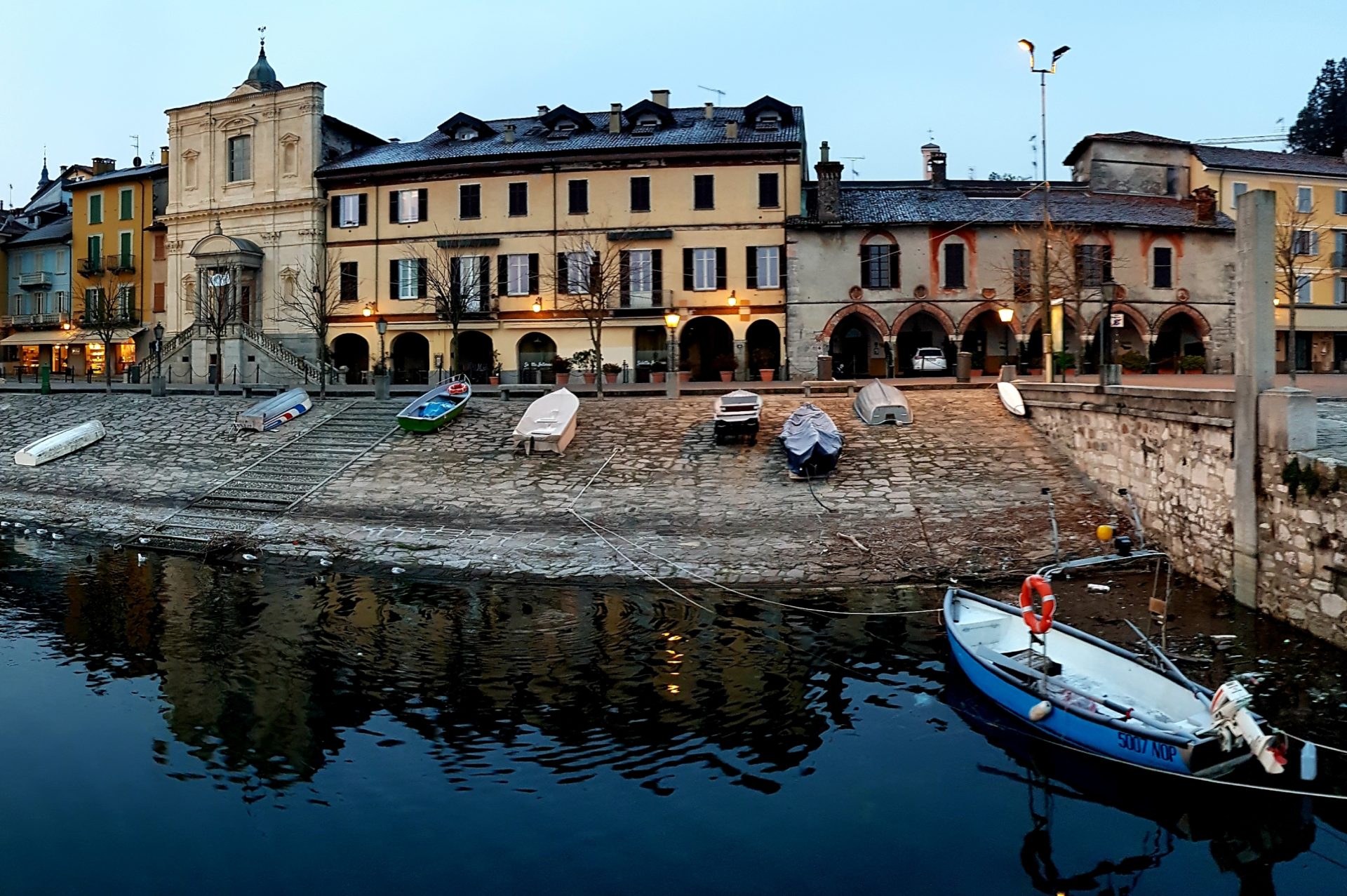 Vista di un porto dall'acqua, all'imbrunire. Una piccola imbarcazione è in acqua, mentre le altre sono tirate a riva, davanti a un edificio giallo illuminato