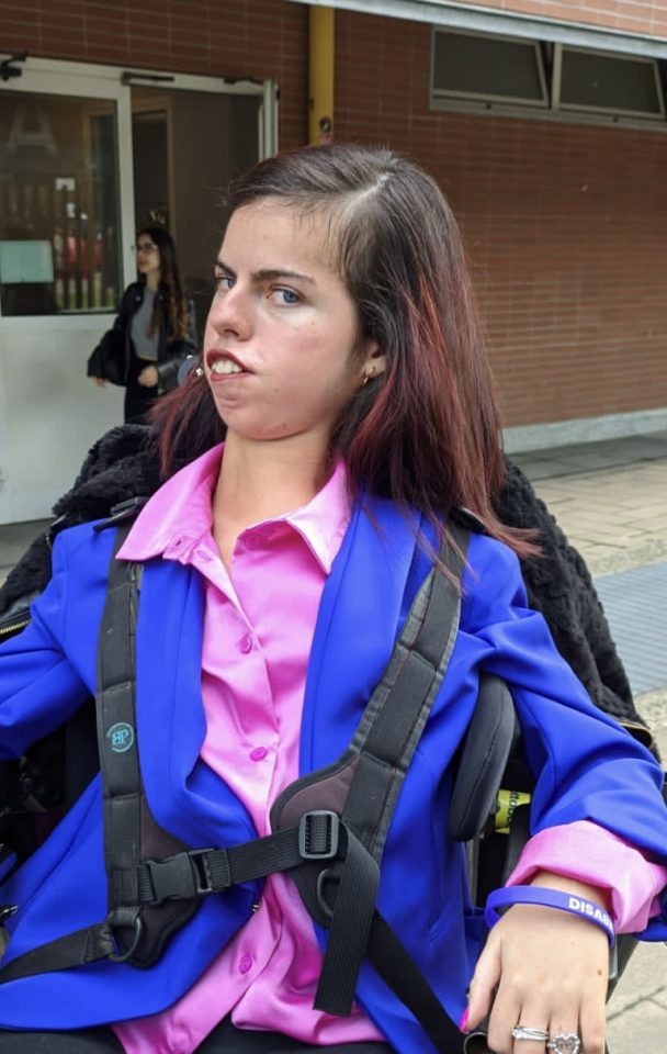 Elisa Costantino seduta su una carrozzina, all'esterno di un edificio, con una camicia rosa e una giacca blu