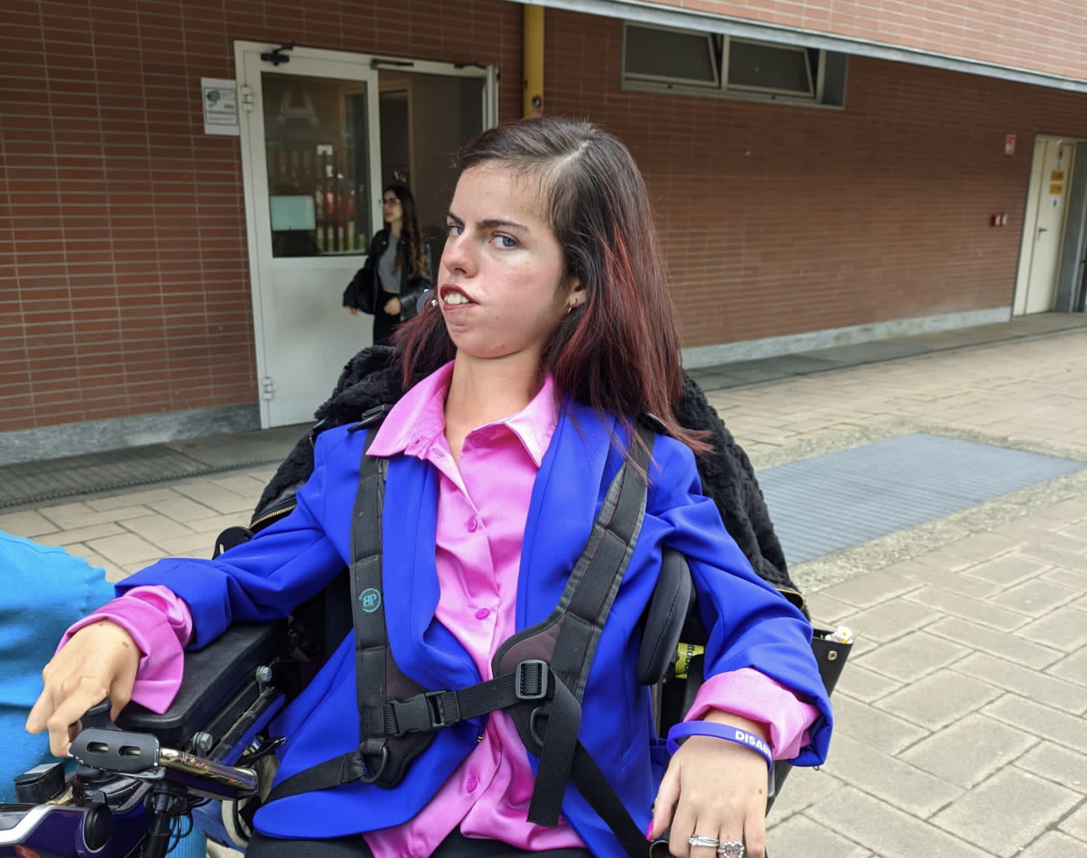 Elisa Costantino seduta su una carrozzina, all'esterno di un edificio, con una camicia rosa e una giacca blu
