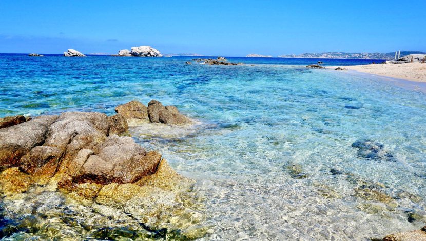 Mare molto azzurro e trasparente, a destra ci sono due scogli, sullo sfondo una barca