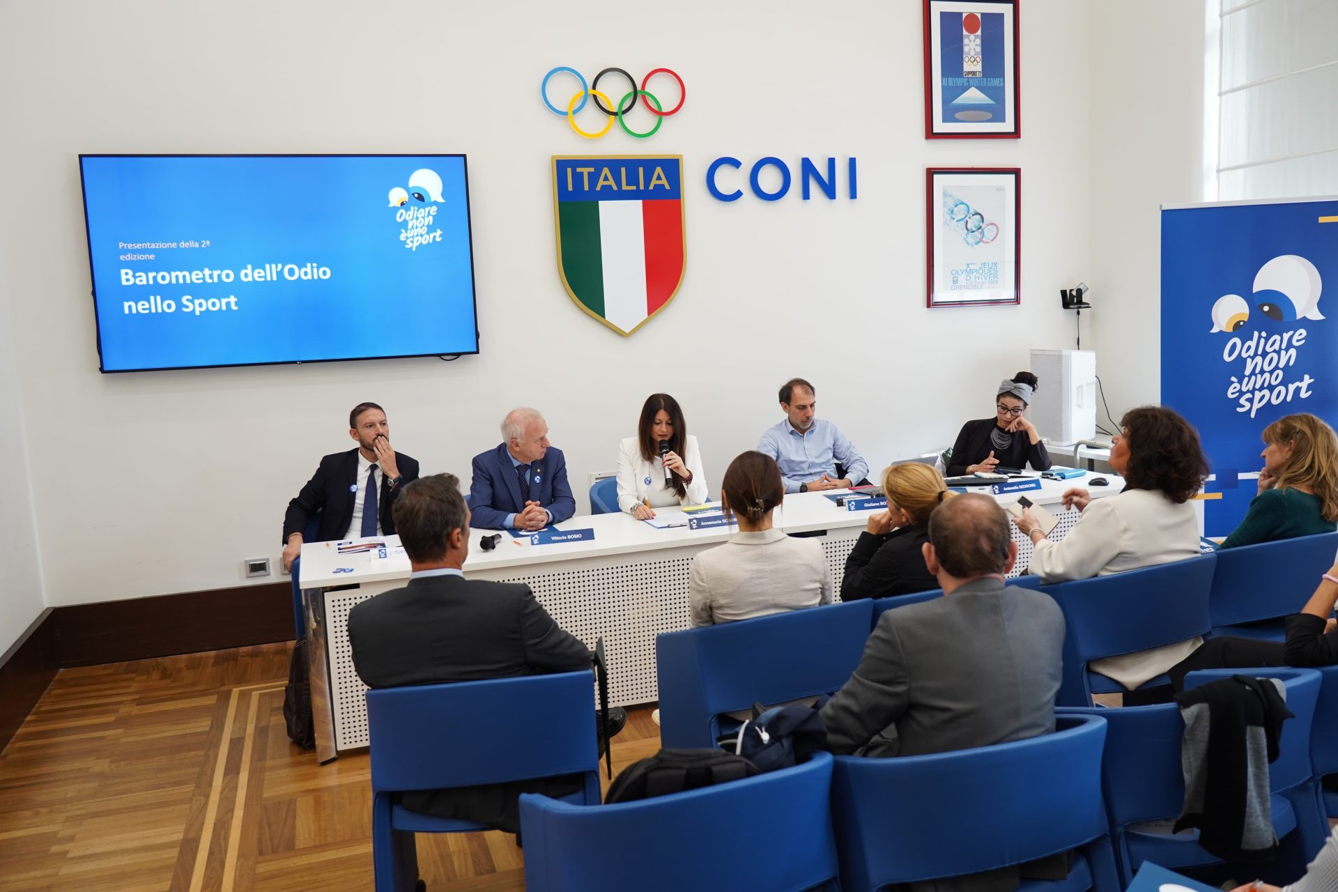 Immagine idi una conferenza, tavolo dei relatori con cinque posti, sul muro lo stemma del Coni e uno schermo con la scritta "Barometro dell'odio nello sport"