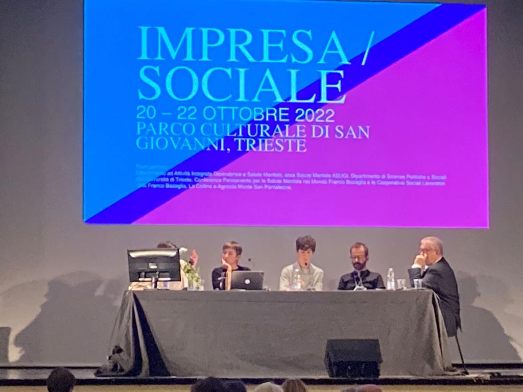 Cinque persone sedute in un tavolo da relatori, sopra uno schermo con scritto "Impresa/sociale"