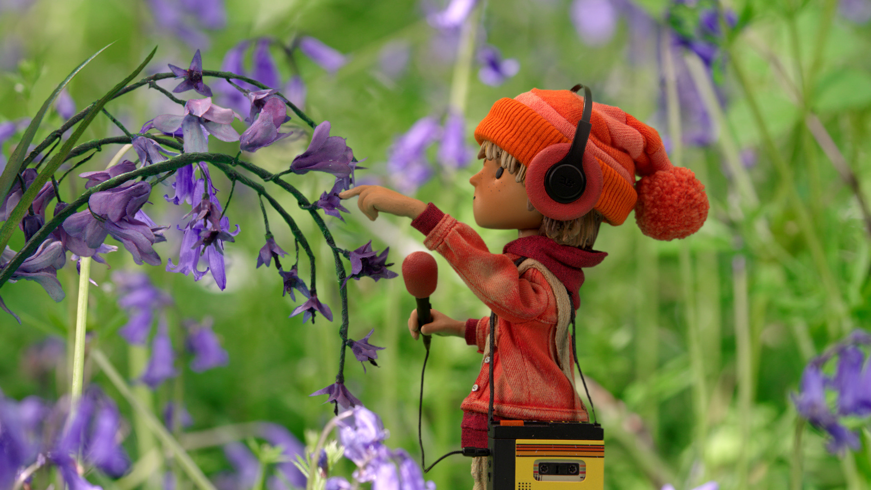 Il cercasuoni vestito d'arancione con le cuffiette e un microfono davanti a dei fiori viola 