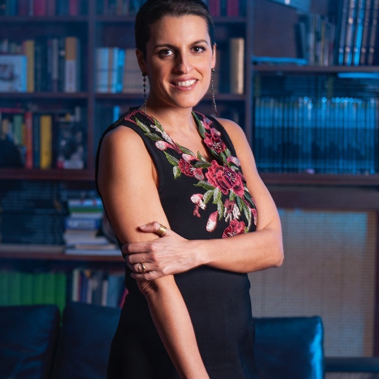 Licia Troisi con un vestito smanicato nero con decorazioni attorno alla scollatura. Ha i capelli neri e corti e sorride. Dietro una libreria in penombra