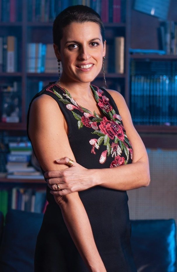 Licia Troisi con un vestito smanicato nero con decorazioni attorno alla scollatura. Ha i capelli neri e corti e sorride. Dietro una libreria in penombra