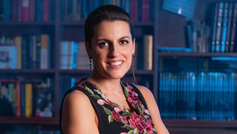 Licia Troisi con un vestito smanicato nero con decorazioni attorno alla scollatura. Ha i capelli neri e corti e sorride. Dietro una libreria in penombra