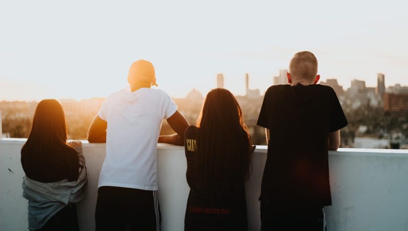 Quattro ragazzi di spalle, appoggiati coi gomiti su una balaustra al tramonto