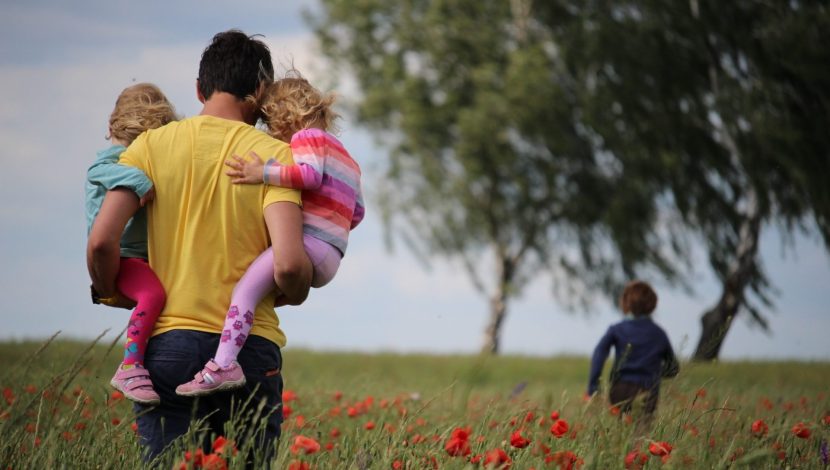 Un padre di spalle con in braccio due bambini in un campo di papaveri, un altro bimbo più avanti, sullo sfondo un albero