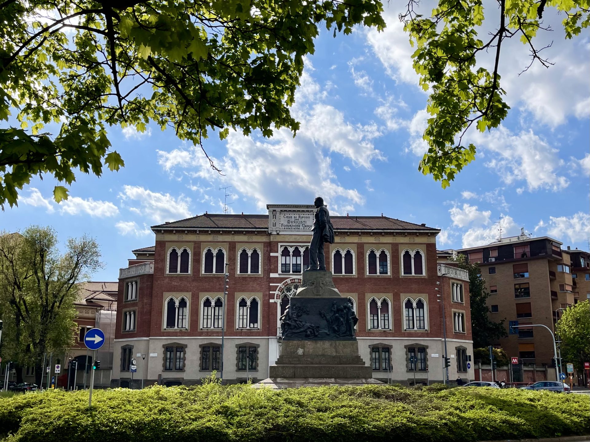 Una villa storica a tre piani in un giorno di sole, davanti una statua e in primissimo piano i rami di un albero