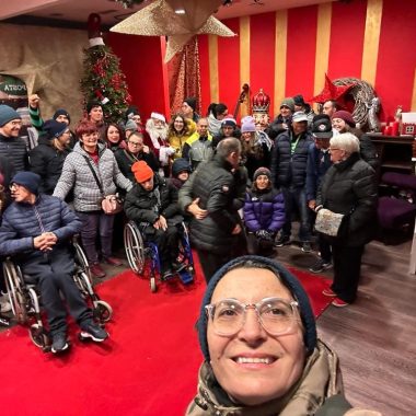 Selfie scattato in una stanza rossa, in primo piano il viso di una donna con gli occhiali, sullo sfondo un gruppo di persone, alcune in carrozzina, altre no