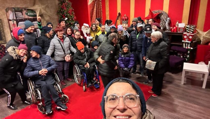 Selfie scattato in una stanza rossa, in primo piano il viso di una donna con gli occhiali, sullo sfondo un gruppo di persone, alcune in carrozzina, altre no