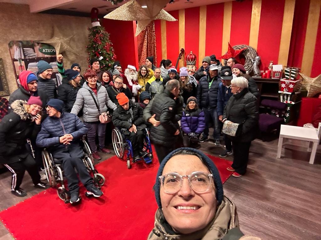 Selfie scattato in una stanza rossa, in primo piano il viso di una donna con gli occhiali, sullo sfondo un gruppo di persone, alcune in carrozzina, altre no