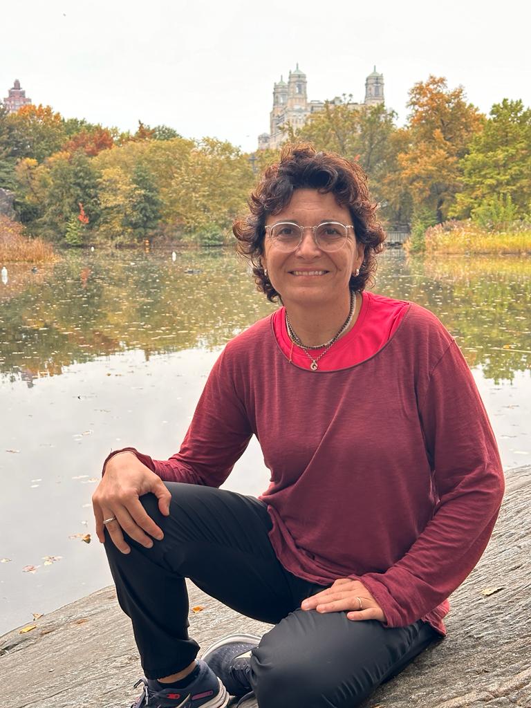 Giuseppina Garritano inginocchiata in riva a un lago, ha i capelli corti e ricci e gli occhiali, porta una maglia fucsia