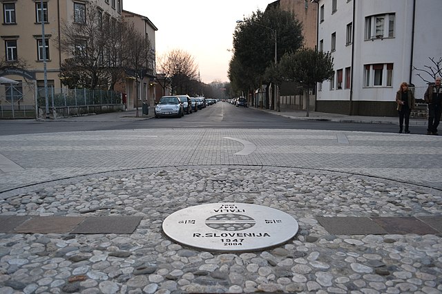 Foto della pavimentazione di una piazza, con al centro una placca tonda con scritto da una parte Italia edall'altra Slovenia