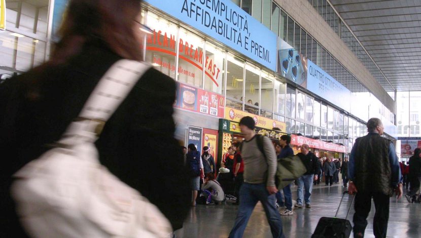 Una stazione affollata, con persone con bagagli che si muovono, in primo piano, di spalle e sfuocata per il movimento, una persona con una borsa bianca