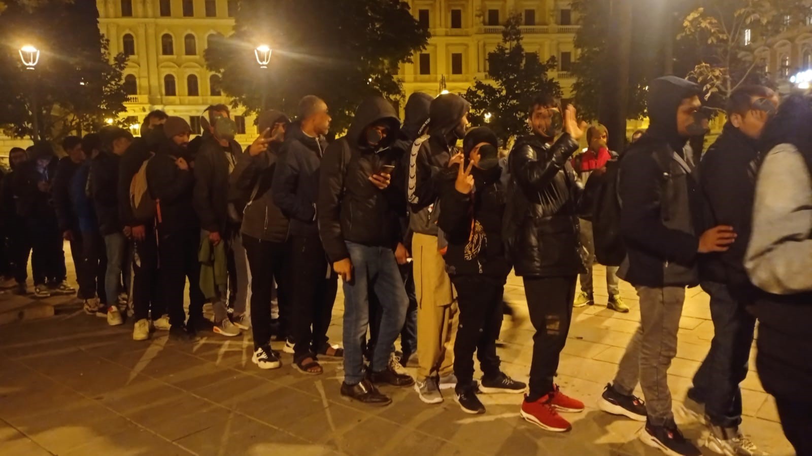 Migranti al buio, in fila, con giacche e vestiti pesanti, in una piazza cittadina