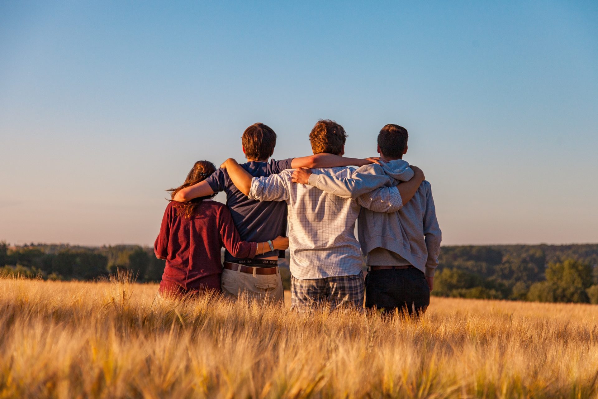 Quattro giovani di spalle abbracciati in un campo di grado, mentre guardano il cielo al tramonto