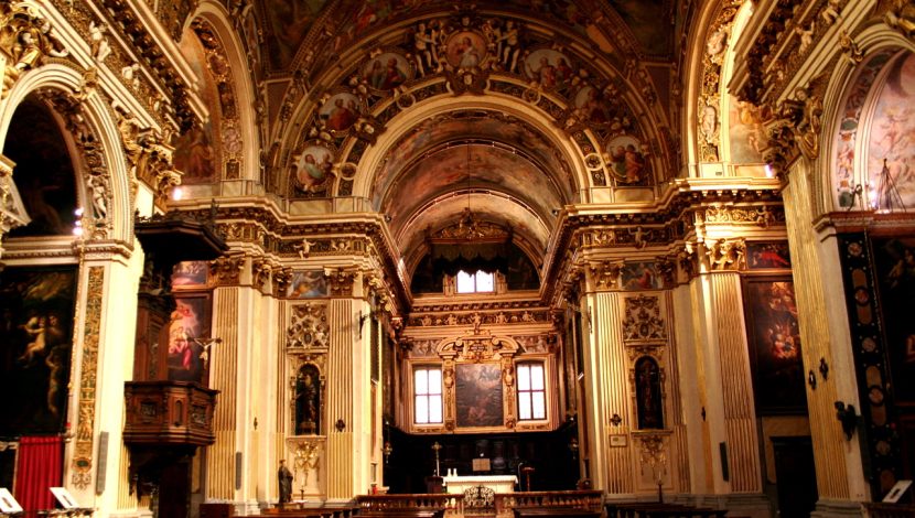 L'interno di una chiesa con prospettiva centrale, di fronte l'abside con l'altare, sulle pareti ci sono degli affreschi