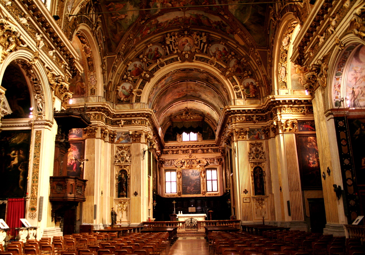 L'interno di una chiesa con prospettiva centrale, di fronte l'abside con l'altare, sulle pareti ci sono degli affreschi