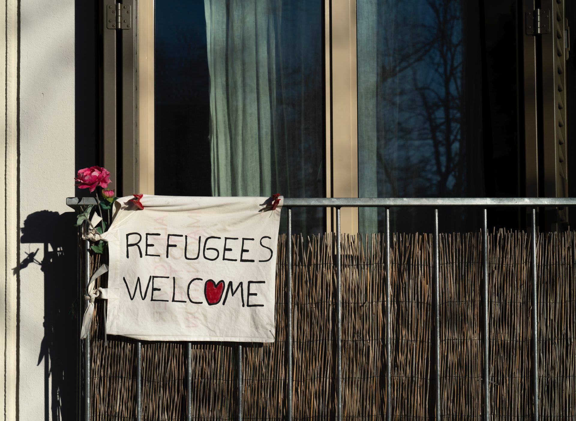 Un balcone con a sinistra appeso alla ringhiera un cartello "refugees welcome", con un cuore al posto della o e una rosa appesa vicino