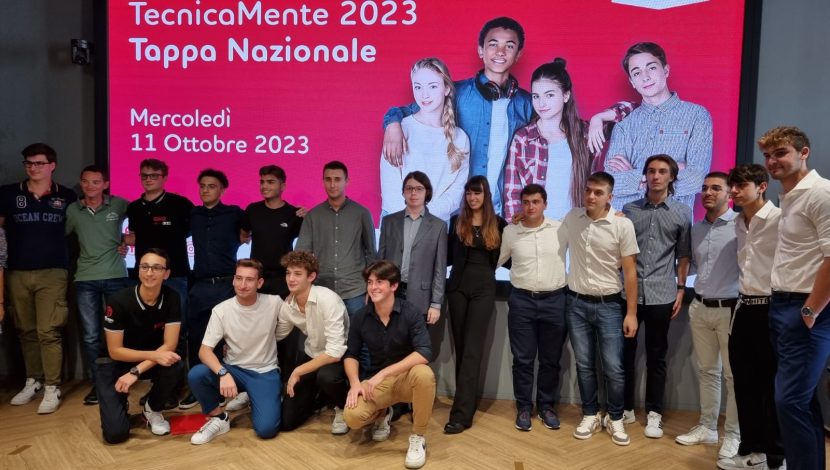 Una classe di ragazzi in posa davanti a un megaschermo con scritto "Tecnicamente 2023, Tappa Nazionale" rosso, l'immagine di quattro ragazzi