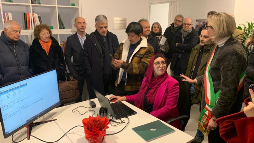 Persone riunite attorno a un tavolo da ufficio, con sulla sinistra il monitor di un computer, a destra una persona con una fascia tricolore