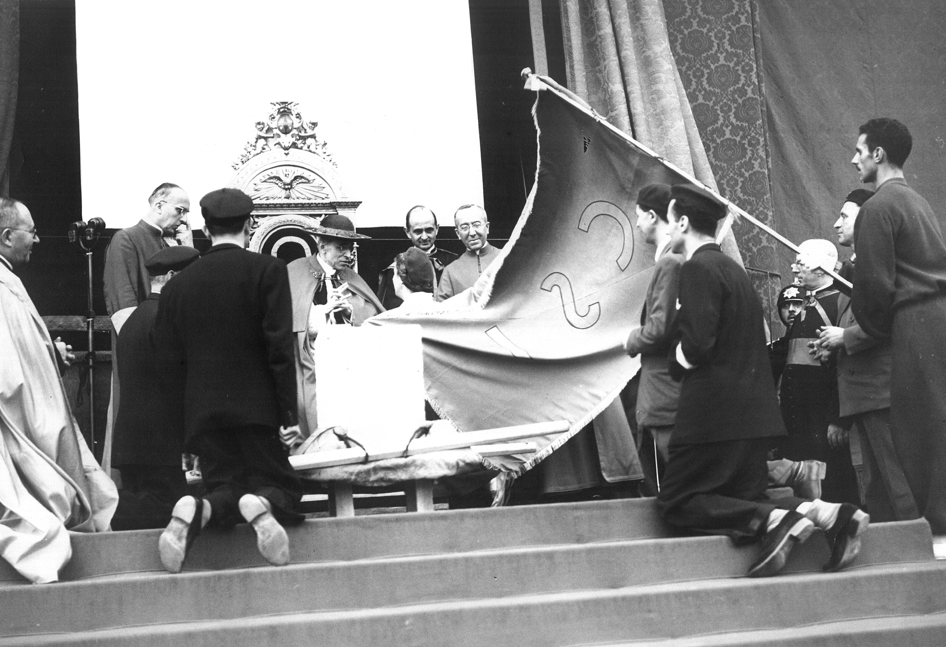 Una foto d'epoca, in bianco e nero. Una grande bandiera entra dalla destra verso il papa seduto. Sulla sinistra, un uomo inginocchiato 