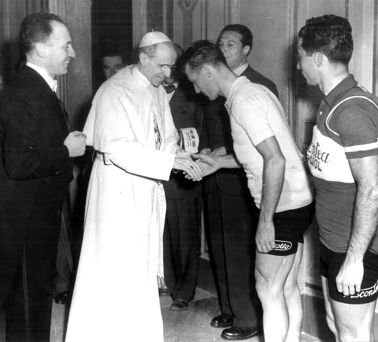 Foto d'epoca in bianco e nero. Al centro il papa vestito di bianco che stringe la mano a una persona vestita con pantaloncini corti e maglietta. Attorno, altre persone 
