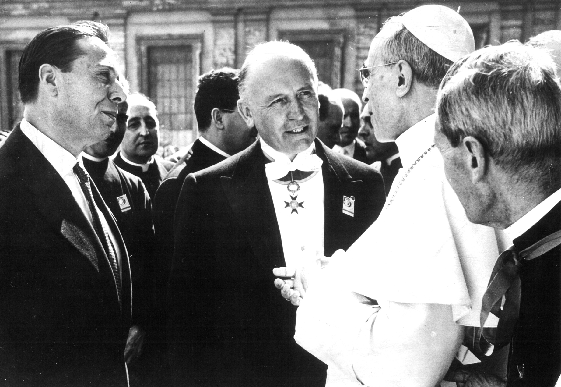 Tre uomini in primo piano, sulla destra il papa vestito di bianco, al centro un uomo con una medaglia al petto, sulla sinistra un altro signore 