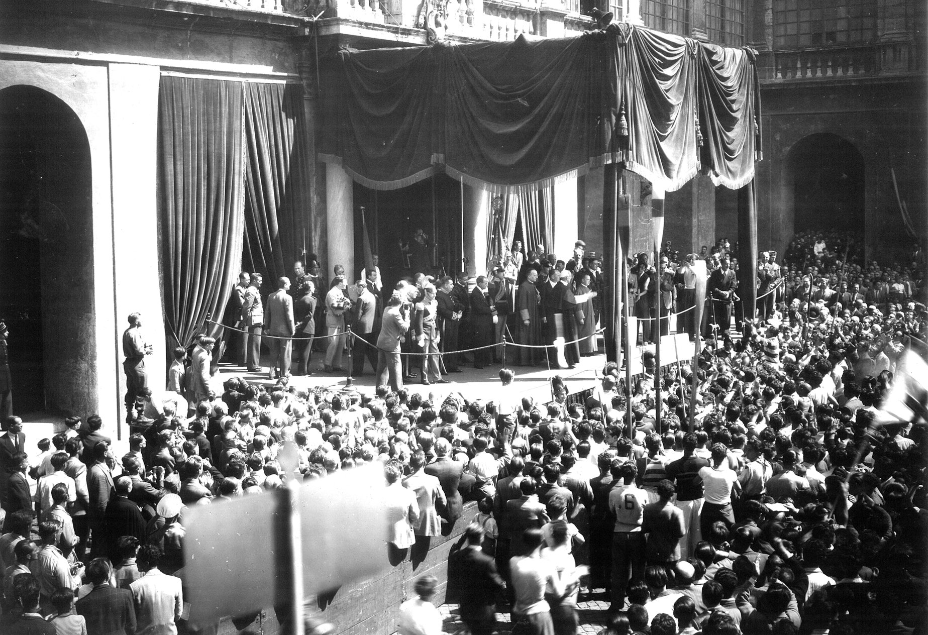 Foto d'epoca in bianco e nero. Un grande baldacchino al centro, con molte persone. Sotto, una grande folla radunata 