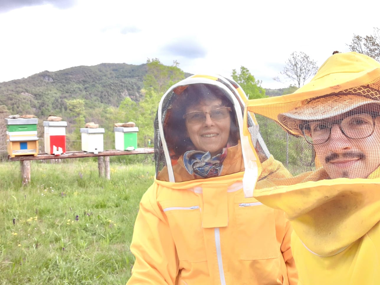 Due persone vestite con completi da apicultori, sullo sfondo prati, arnie e in lontananza una montagna 