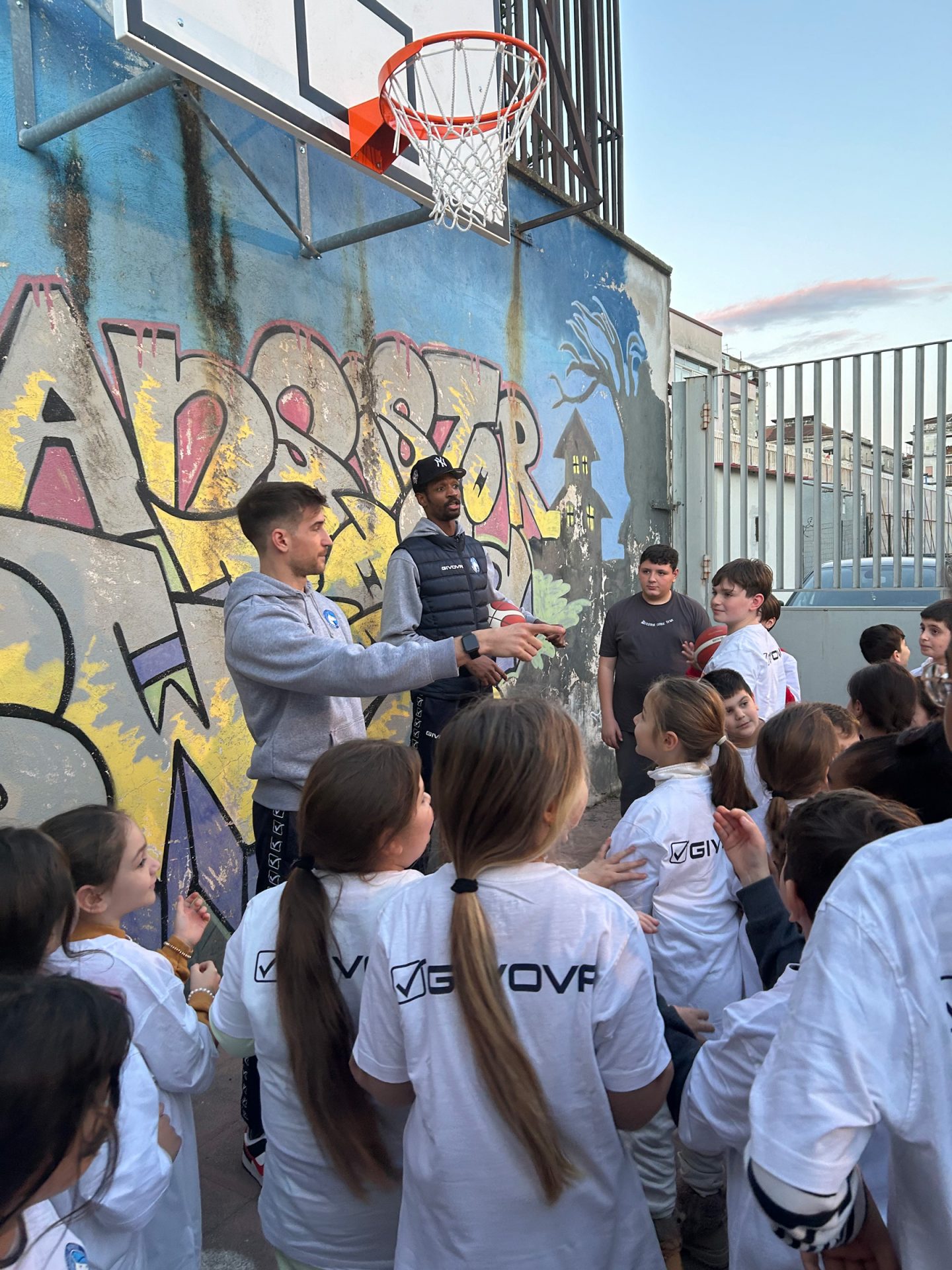 Una folla di bambini di spalle e due giocatori davanti a un muro con un graffito, sotto a un canestro 