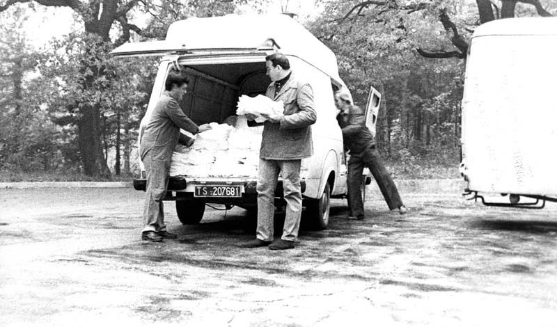Foto d'epoca in bianco e nero, due persone che caricano degli asciugamani su un furgone, un'altra persona che entra dal sedile del passeggero