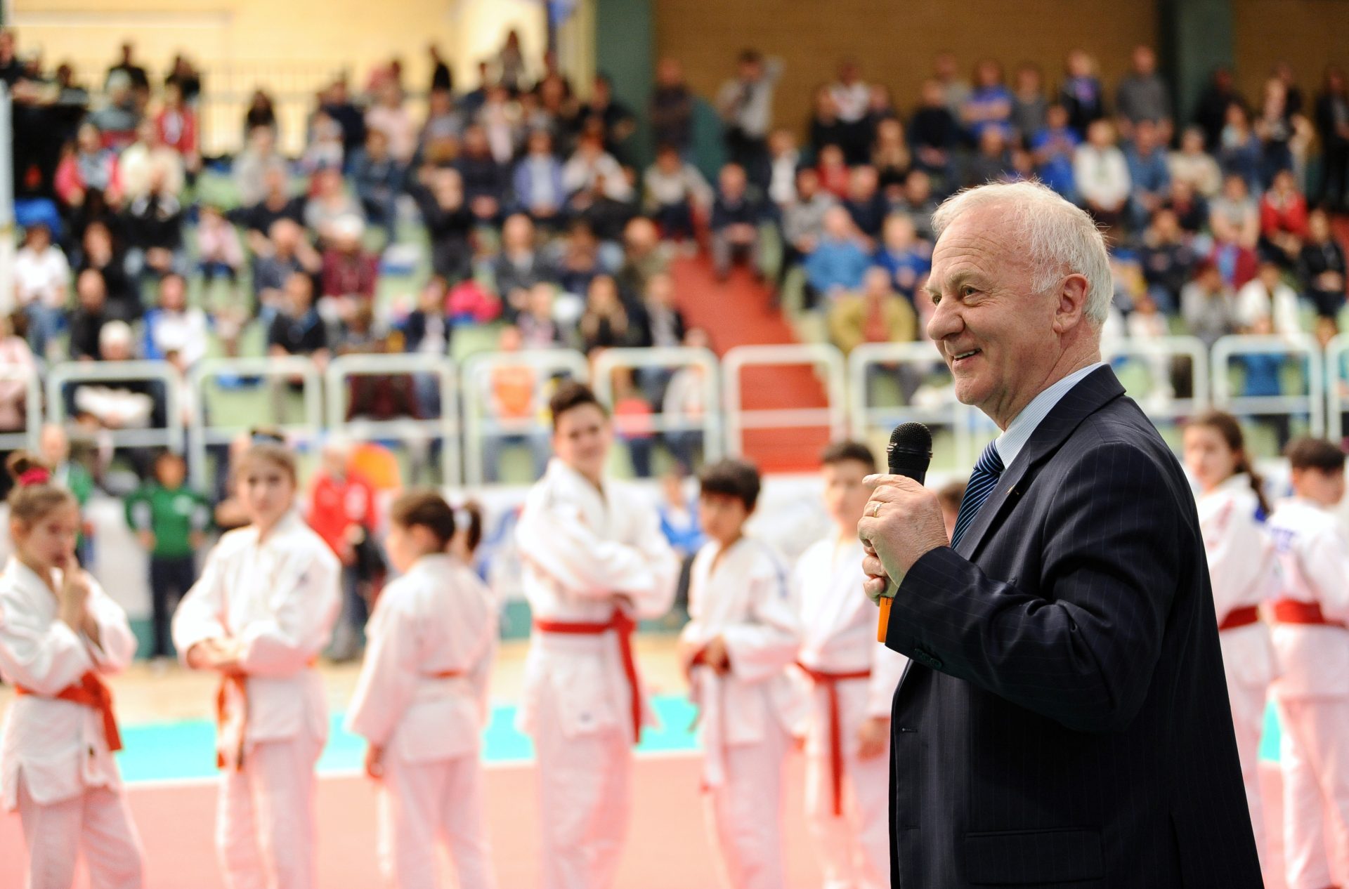 Un uomo in giacca e cravatta sulla destra, sullo sfondo un palazzetto con alcuni ragazzi vestiti con judoji e persone sugli spalti 