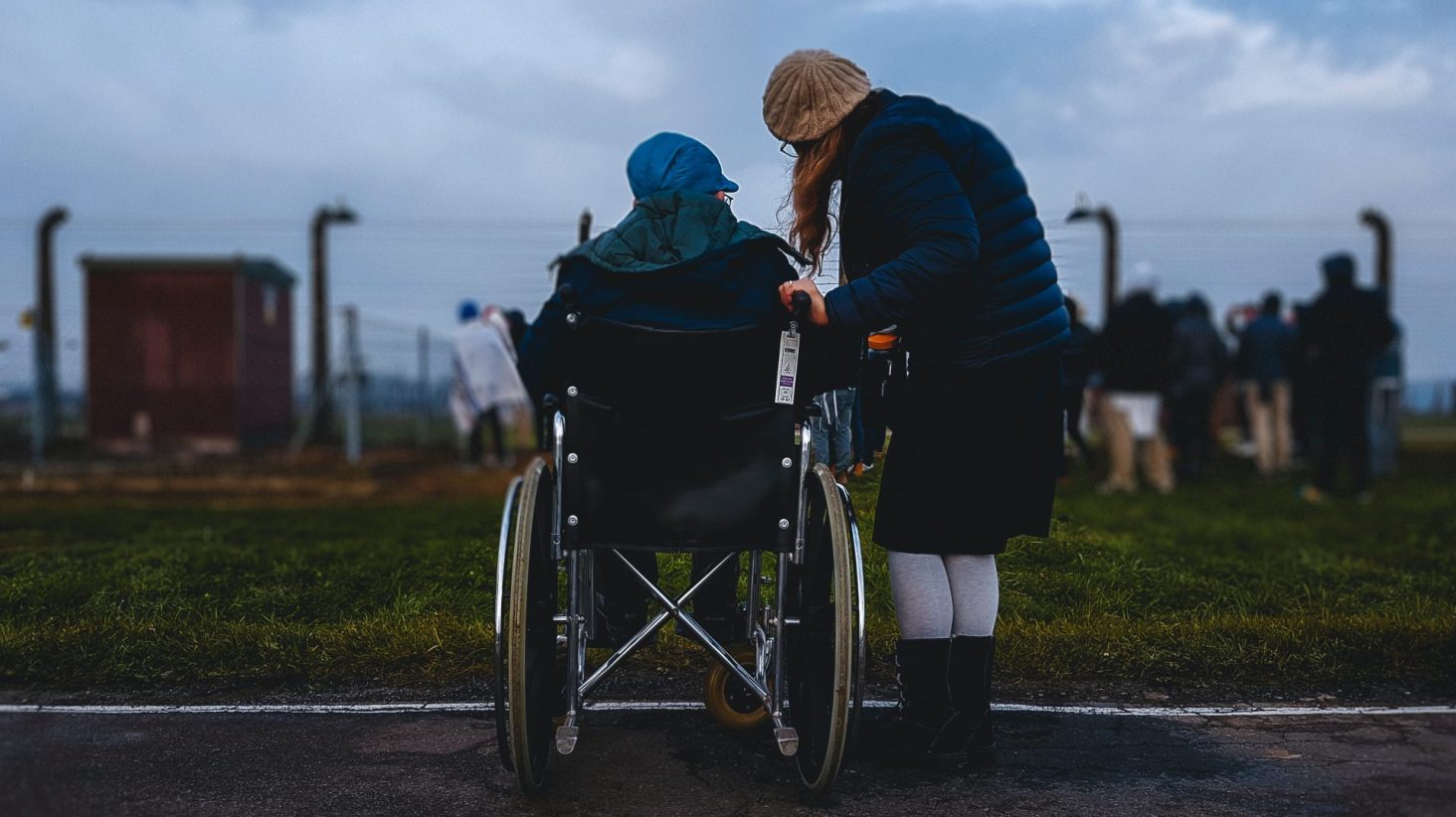 Una persona in sedia a rotelle con vicino una persona in piedi, entrambe di spalle, davanti a un parco cittadino con un cielo scuro