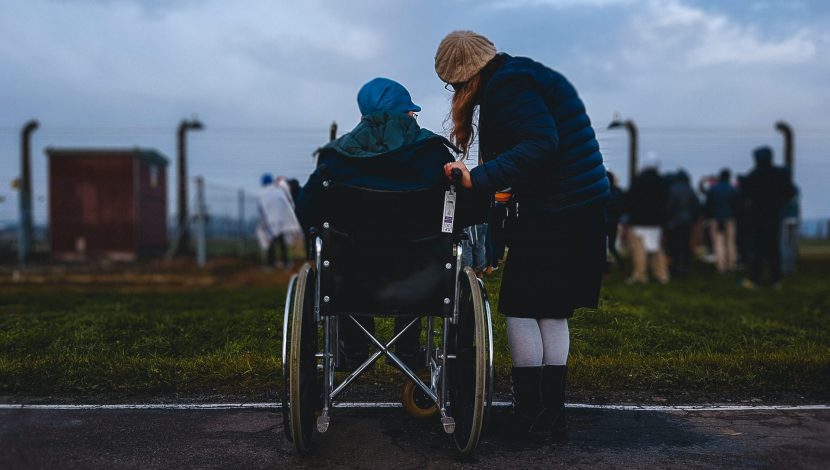 Una persona in sedia a rotelle con vicino una persona in piedi, entrambe di spalle, davanti a un parco cittadino con un cielo scuro