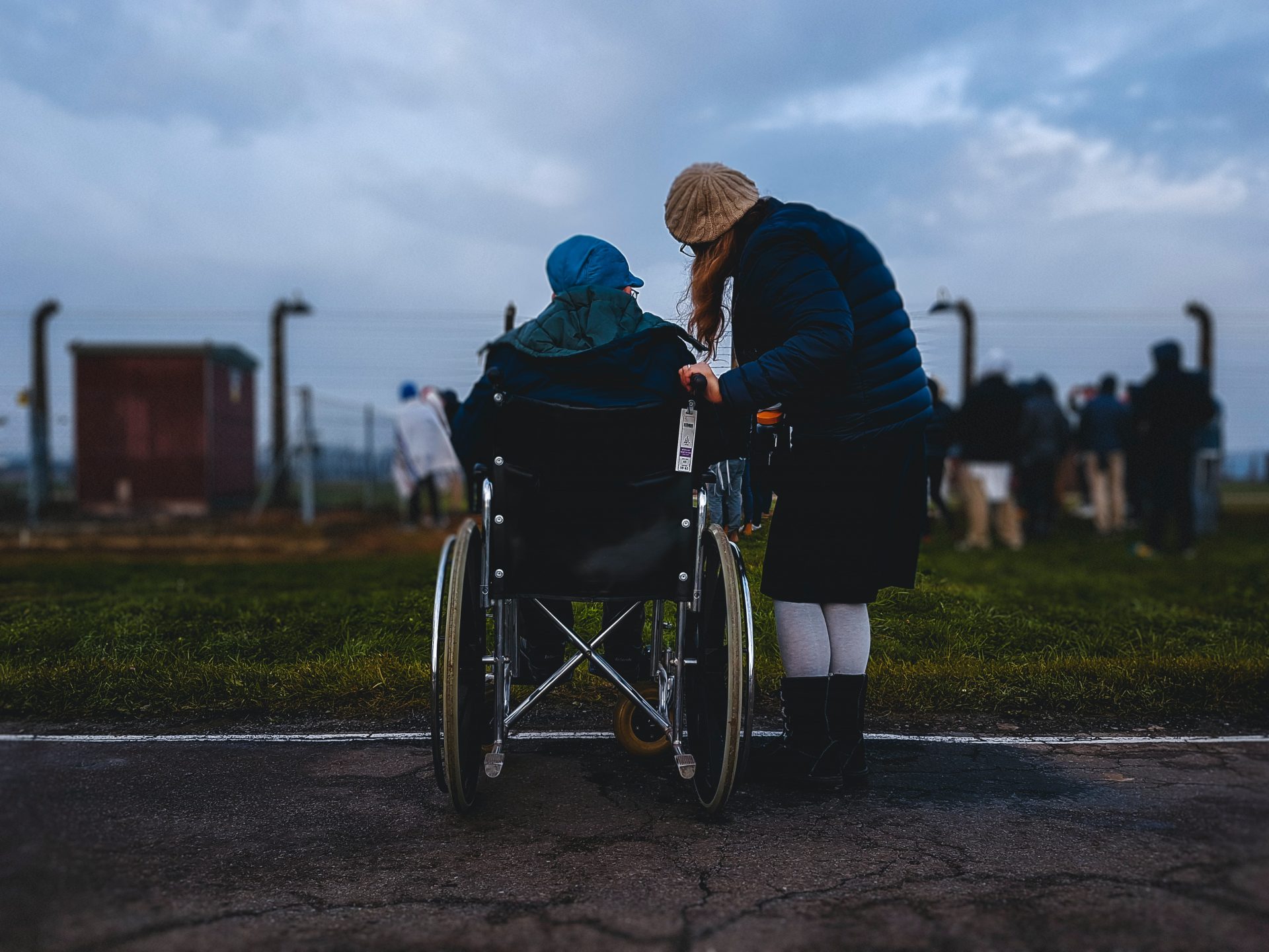 Una persona in sedia a rotelle con vicino una persona in piedi, entrambe di spalle, davanti a un parco cittadino con un cielo scuro