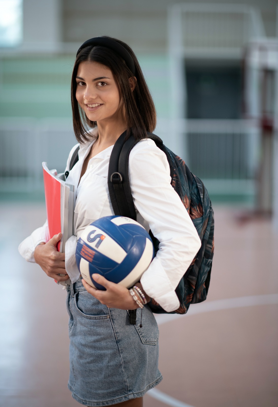 Una ragazza con uno zaino sulle spalle, in mano un quaderno e una palla da pallavolo