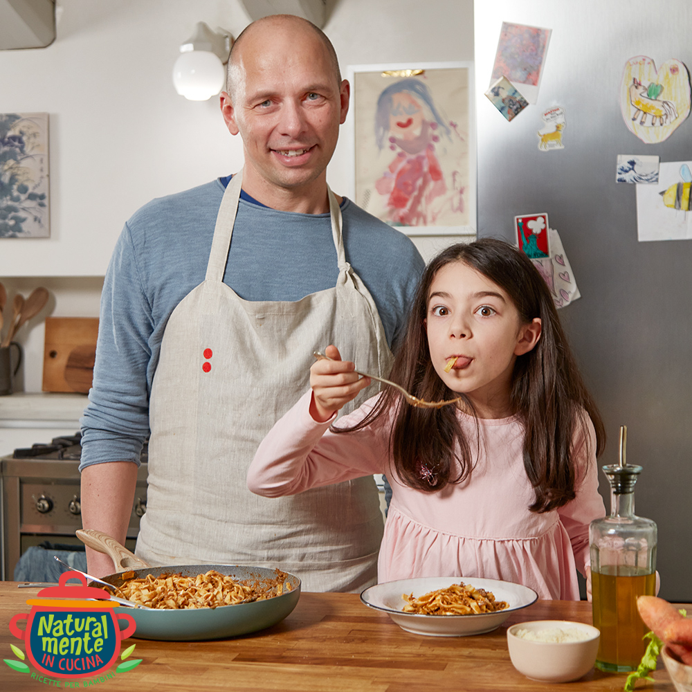 Un uomo e una bambina dietro a un tavolo da cucina su cui è appoggiata una pentola di tagliatelle. La bambina ne assaggia un po'