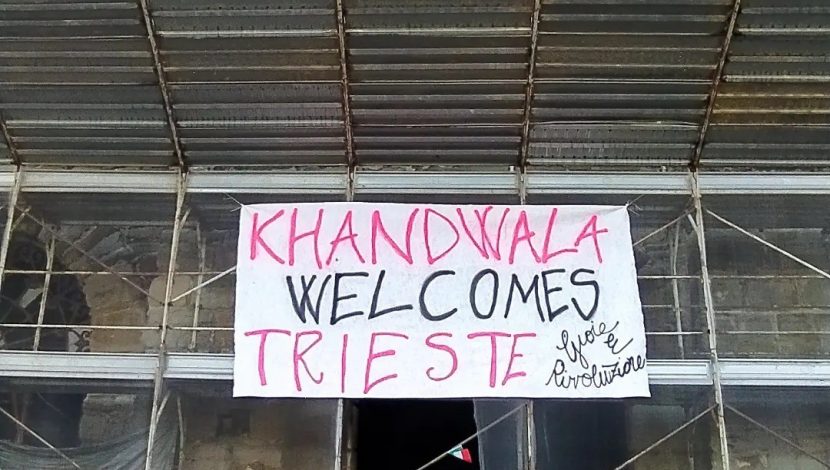 L'ingresso di un edificio coperto di impalcature, con un lenzuolo con la scritta "Khandwala welcomes Trieste"