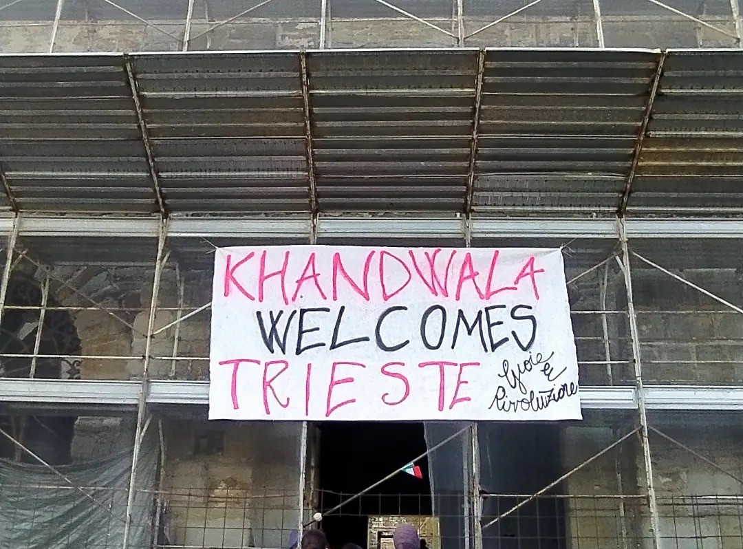 L'ingresso di un edificio coperto di impalcature, con un lenzuolo con la scritta "Khandwala welcomes Trieste"