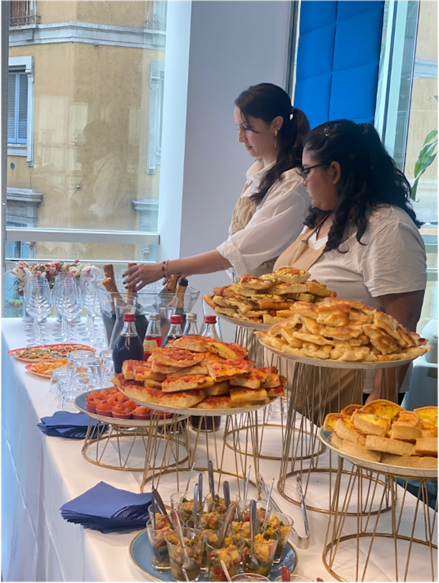 De persone dietro a un tavolo in cui è allestito un catering con pizzette e bottiglie