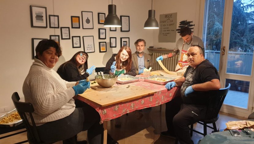 Alcune persone sedute intorno a un tavolo, con dei guanti, stanno lavorando della pasta fresca