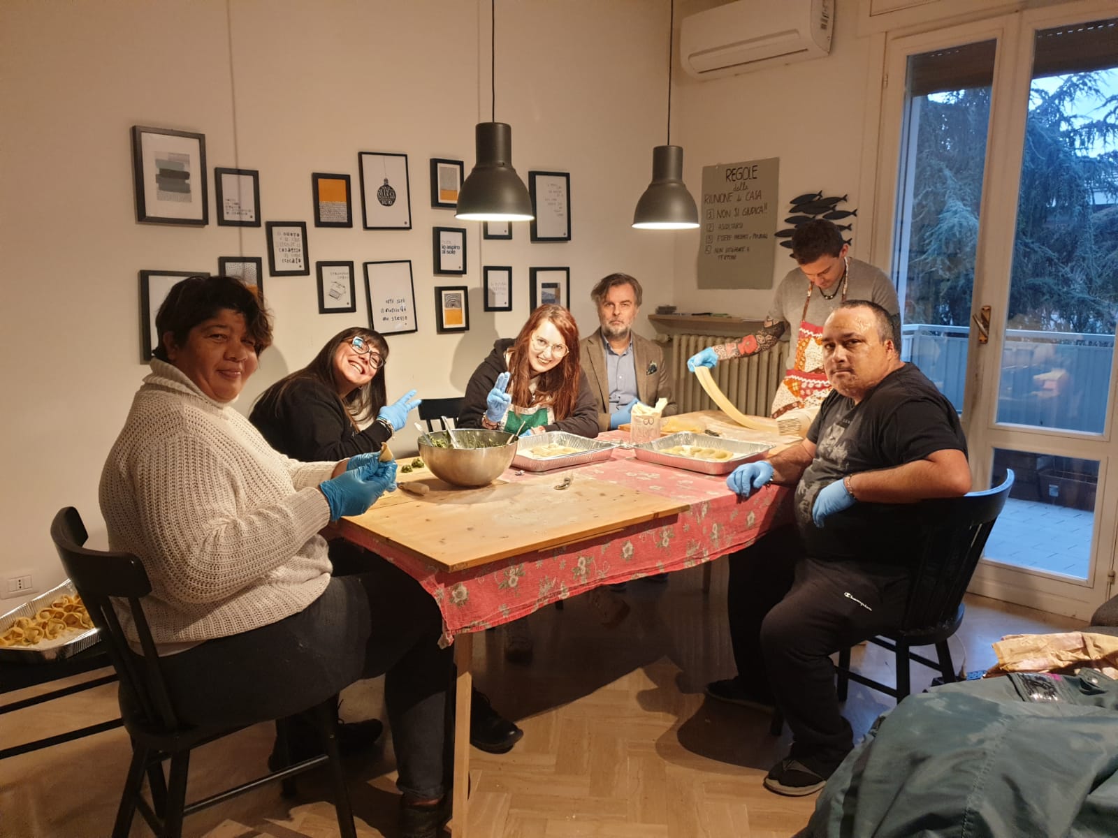 Alcune persone sedute intorno a un tavolo, con dei guanti, stanno lavorando della pasta fresca