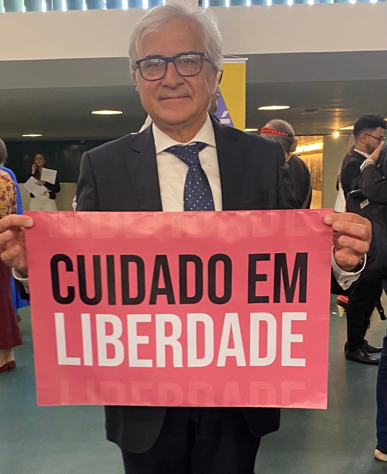 Roberto Mezzina in giacca e cravatta, con un cartello su cui è scritto "Cuidado em liberdade"