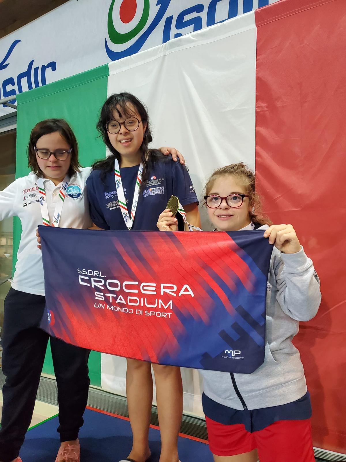 Tre ragazze su un podio, tengono in mano una bandiera con scritto "Crocera stadium"
